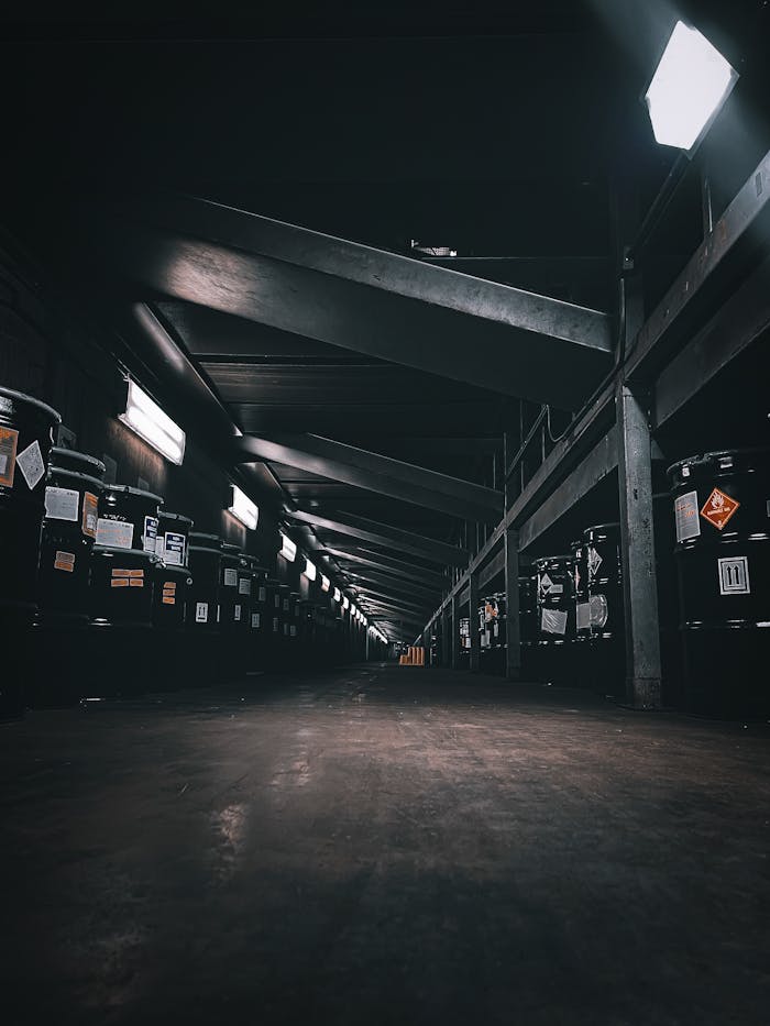 A dark, illuminated warehouse corridor with rows of storage barrels and beams overhead.