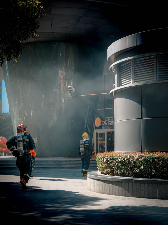Firefighters in action outside a modern urban building, smoke visible, wearing safety gear.